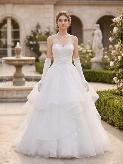 A woman wearing a strapless white wedding gown with a fitted lace bodice, long white gloves, and a full layered tulle skirt, standing in a garden courtyard with a fountain and stone building in the background.