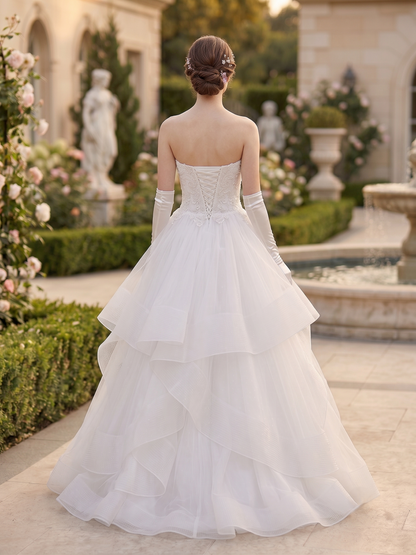 A woman stands outdoors in a grand garden wearing a strapless white wedding gown with long gloves and a full layered tulle skirt. The background shows statues, flowers, hedges, and a fountain.