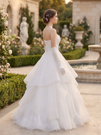 A woman wearing a white strapless wedding gown with long white gloves, photographed outdoors in a garden courtyard with statues, flowers, and a fountain in the background.