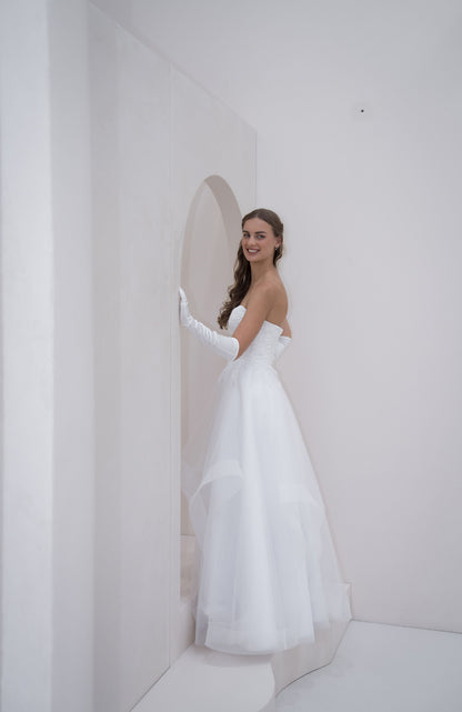 Woman in a white wedding dress standing in a minimalistic room with a white wall and floor.