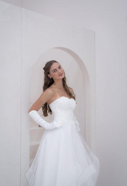 Woman in a white strapless gown with long gloves standing against a white wall.