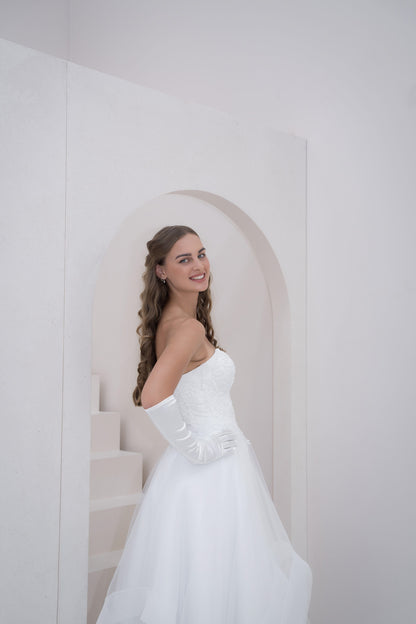 Woman in a white wedding dress standing in a softly lit room with a white wall and archway.