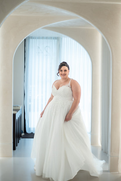 Woman in a white wedding dress standing in an archway with light streaming through curtains.