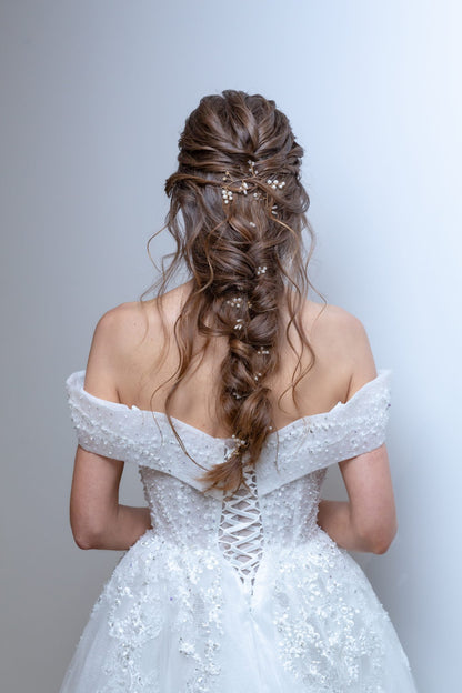 Woman in a white wedding dress with intricate detailing, standing against a plain background.