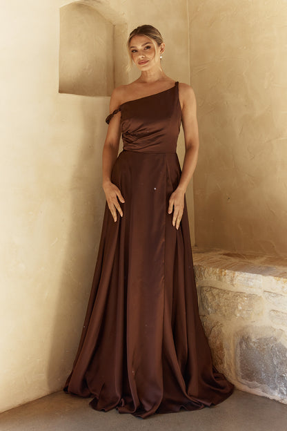 Woman in a brown one-shoulder gown standing against a neutral wall.