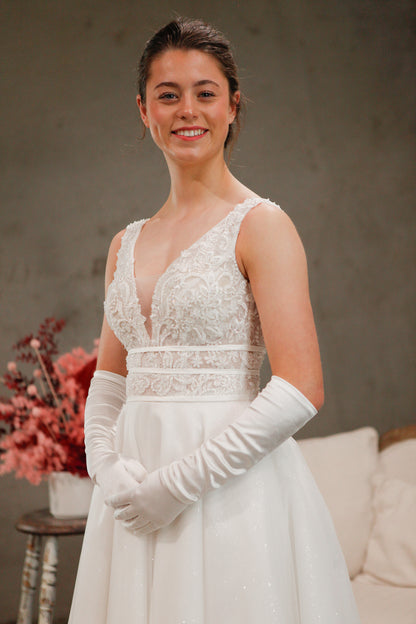 Woman in a white wedding dress with lace detailing and long gloves, standing against a neutral background.
