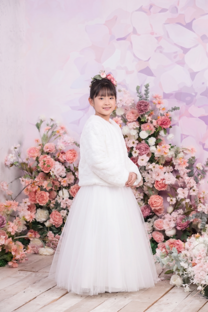 A young girl stands in a white long-sleeve dress with a full tulle skirt, posed in front of a decorative floral arrangement and a soft pink backdrop.