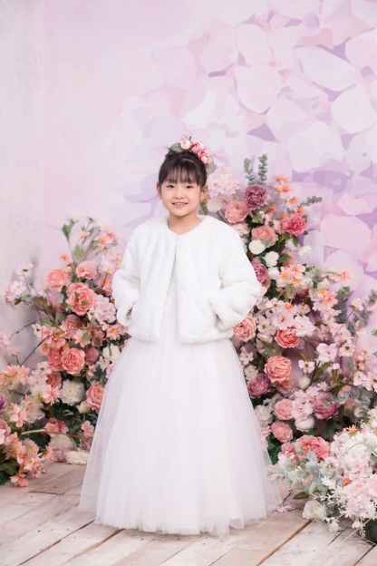 A young girl stands in a white tulle dress with a white faux-fur jacket, posing in front of a pink floral backdrop.
