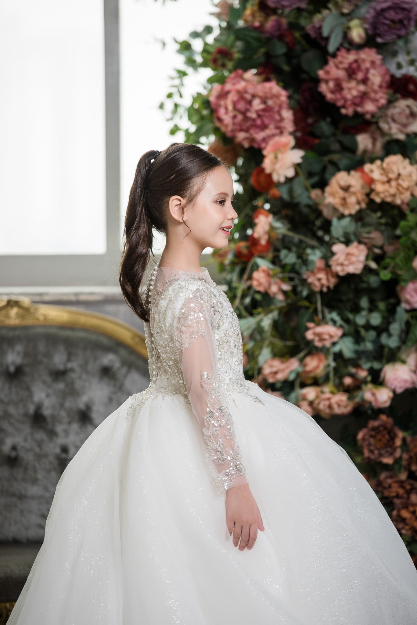 Young girl in a white dress standing in front of a floral arrangement.