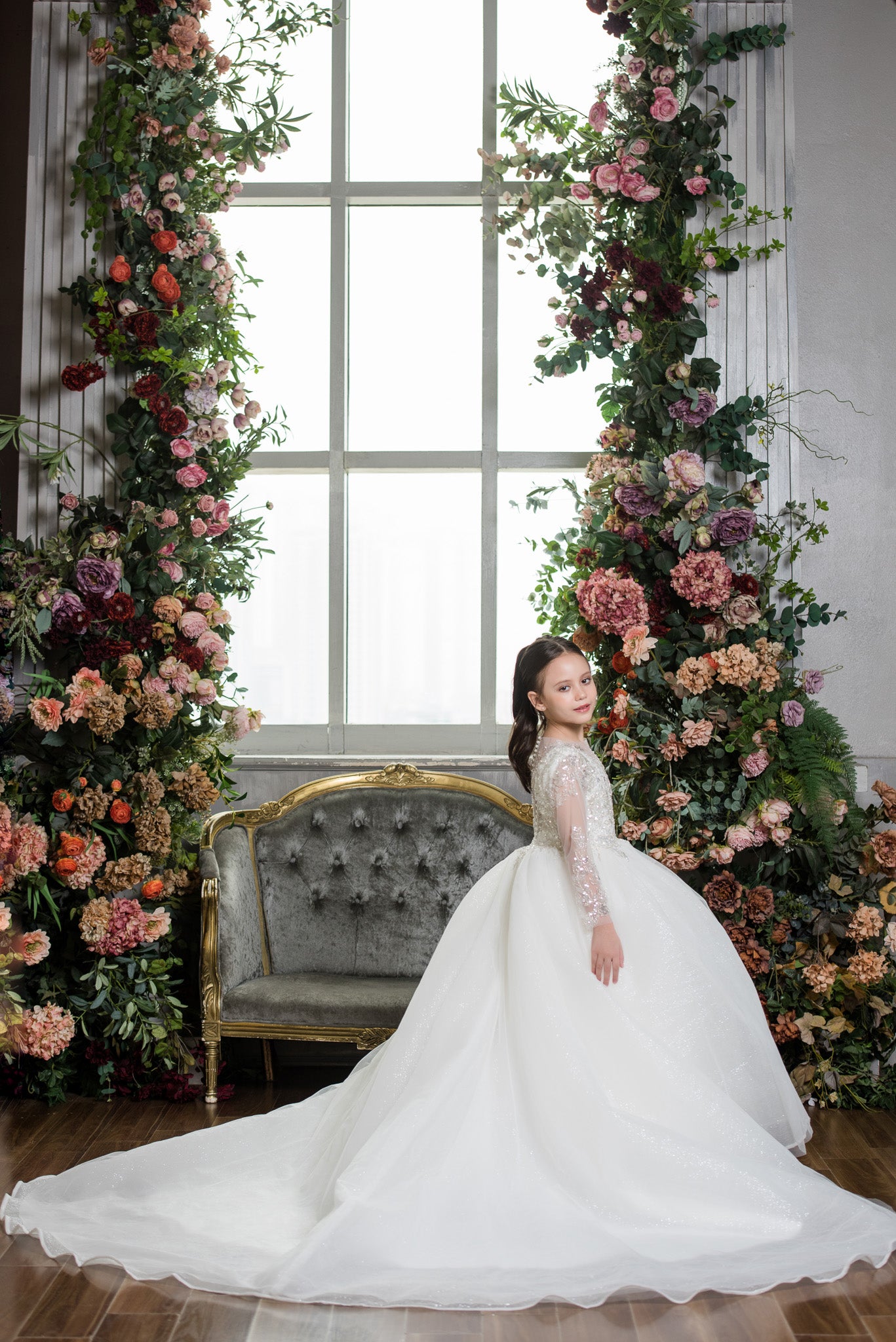 Woman in a white dress standing in front of floral decorations and a window.