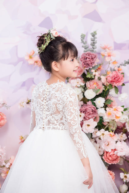 Young girl in a white lace dress standing in front of a floral arrangement with a light purple background