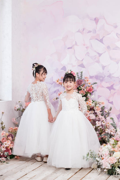 Two young girls in white dresses standing in front of a floral backdrop.
