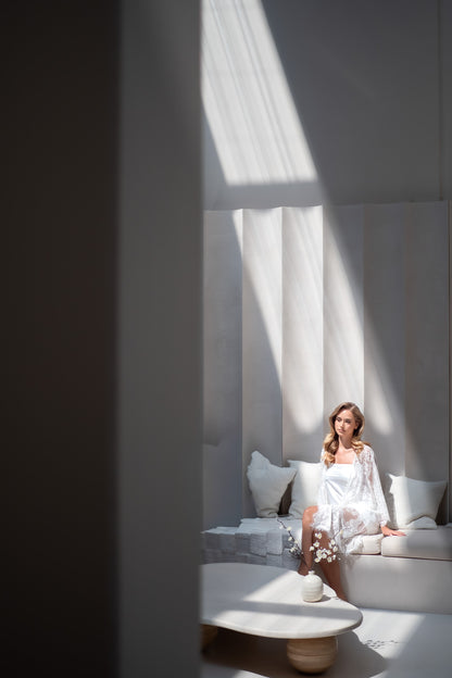 Woman in a white dress sitting on a couch in a bright room with white curtains