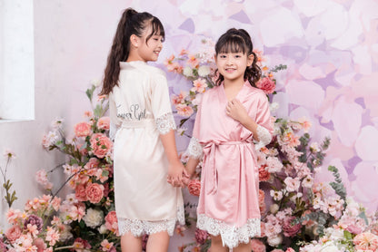 Two children in white and pink robes standing in front of a floral backdrop