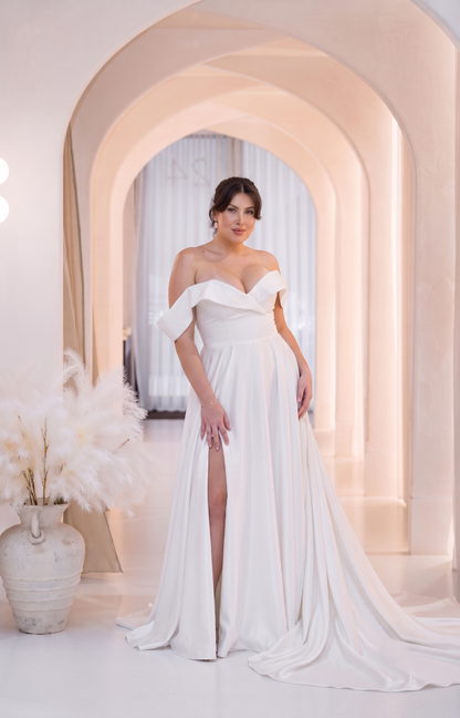 A woman models an elegant off-the-shoulder white wedding gown with a high thigh slit, standing in a softly lit arched hallway with a long train.