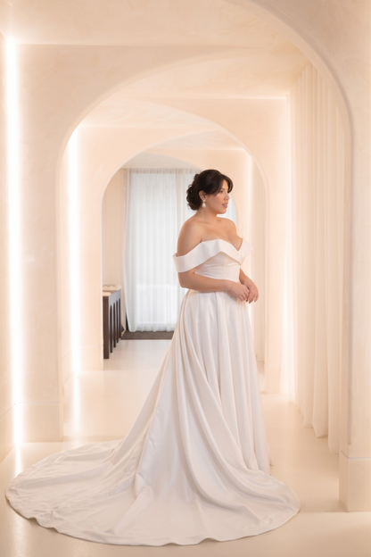 A woman wearing an off-the-shoulder white wedding gown with a long train, standing in a softly lit arched hallway.