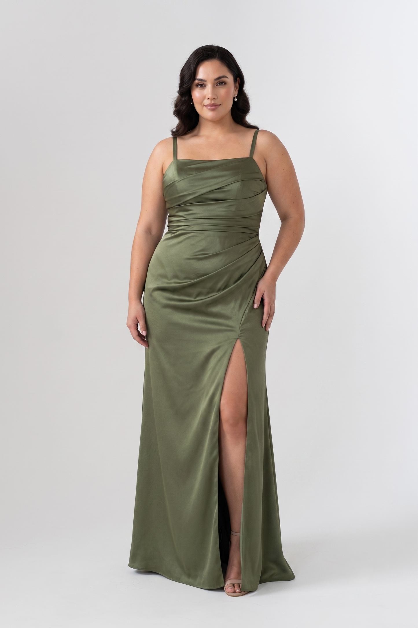 A woman modeling a floor-length olive green satin gown with spaghetti straps, a draped sweetheart neckline, and a high thigh slit, photographed against a light gray studio background.