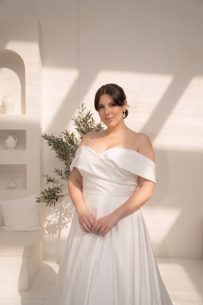 A woman wearing an off-the-shoulder white A-line dress, standing in a bright, minimalist white room with architectural shadows.