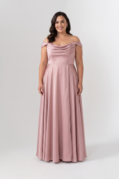 A woman modeling a floor-length off-the-shoulder bridesmaid dress in dusty pink with a draped bodice and flared skirt, photographed against a light gray studio background.