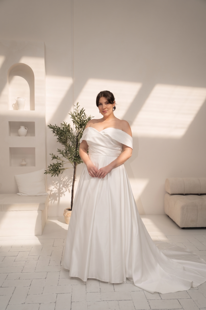 A woman wearing a white off-the-shoulder A-line wedding gown with a long train, standing in a bright, modern white room with architectural shadows.