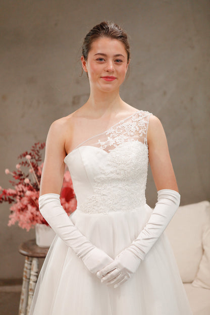 Woman in a white wedding dress with lace detailing and long gloves, standing against a neutral background.