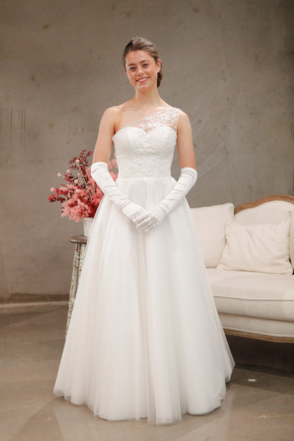 Woman in a white wedding dress with lace detailing and white gloves, holding flowers, in a room with a couch.