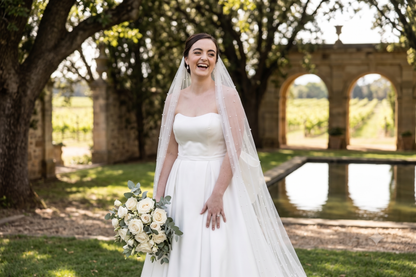 A smiling bride in a strapless white wedding gown with a long sheer veil holds a bouquet of white flowers outdoors near stone arches and a reflecting pool.