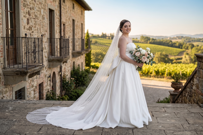 A bride in a strapless white wedding gown with a long sheer veil holds a bouquet of blush and cream flowers, standing outdoors on a stone terrace with a rustic stone building and vineyard landscape in the background.
