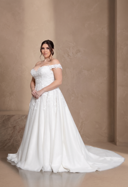 A woman modeling an off-the-shoulder white wedding gown with a fitted lace bodice and a full flowing skirt with a train, posed against a warm beige textured studio backdrop.