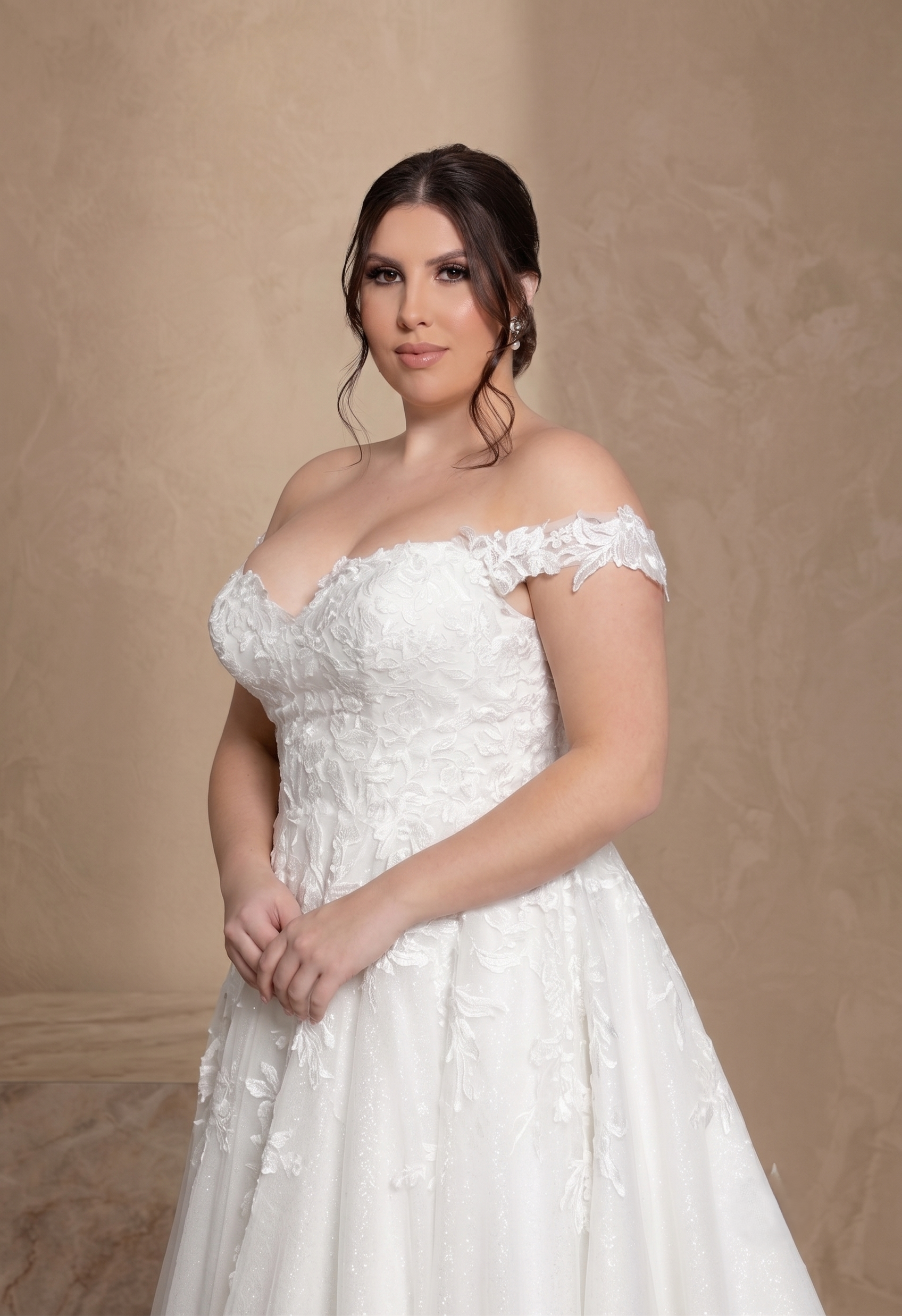 A woman modeling an off-the-shoulder white lace wedding gown against a textured beige studio backdrop.