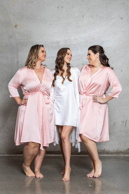 Three women wearing pink and white satin robes against a gray concrete wall.