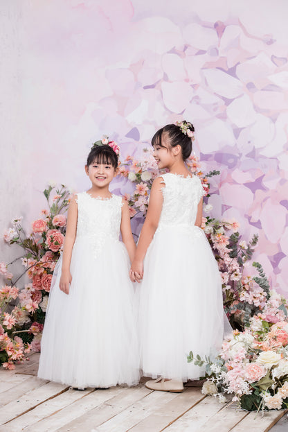 Two young girls in white dresses standing in front of a floral and butterfly backdrop.