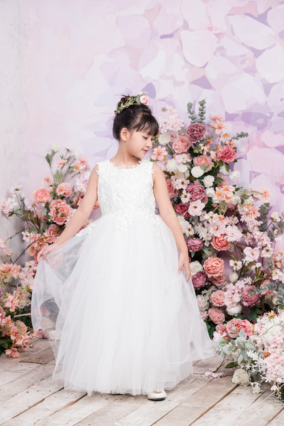 Young girl in a white dress standing in front of a floral arrangement with a soft, blurred background.