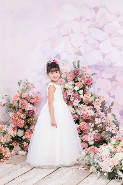 Young girl in a white dress standing in front of a floral arrangement with a soft, pastel-colored background.
