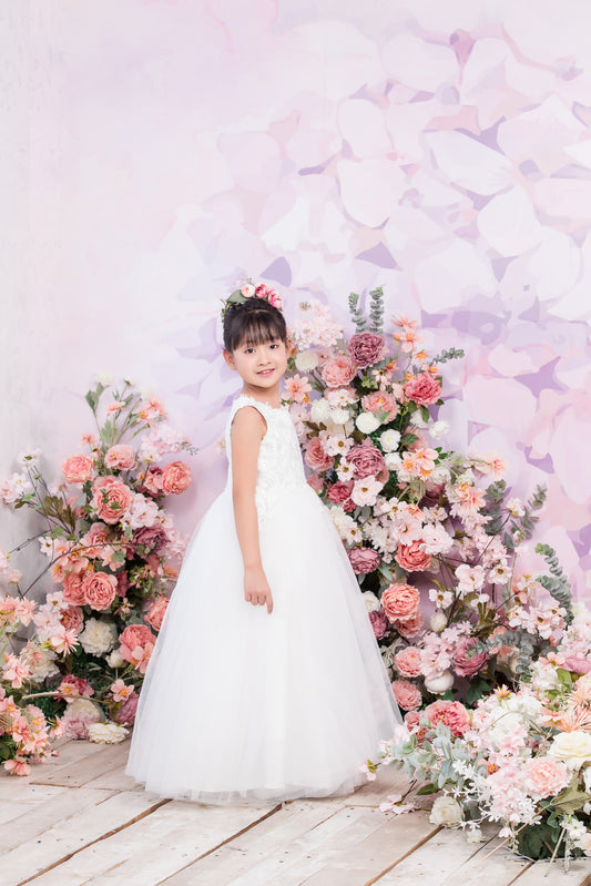 Young girl in a white dress standing in front of a floral arrangement with a soft, pastel-colored background.