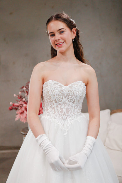 Woman in a white wedding dress with lace detailing and white gloves, standing against a neutral background.