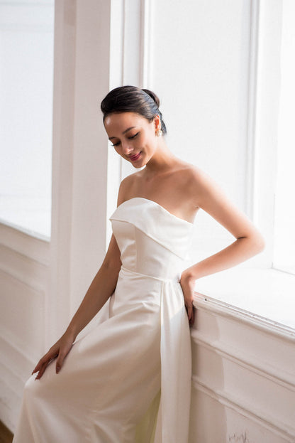 Woman in a white strapless dress standing by a window with bright light.