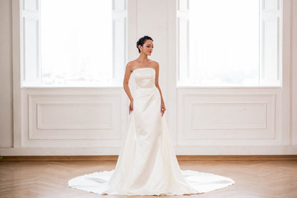 Woman in a white strapless wedding dress standing in a bright room with large windows.