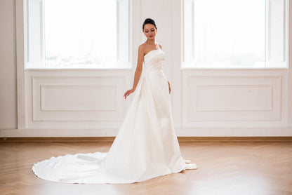 Woman in a white wedding dress standing in a bright room with large windows.