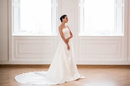 Woman in a white wedding dress standing in a bright room with large windows.