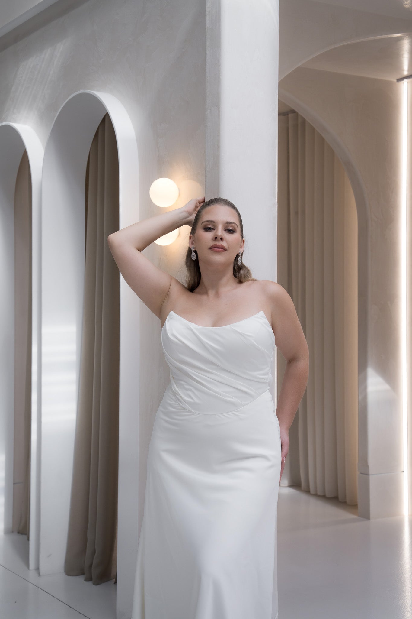 Woman in a white dress standing in a modern architectural setting with arches and soft lighting.
