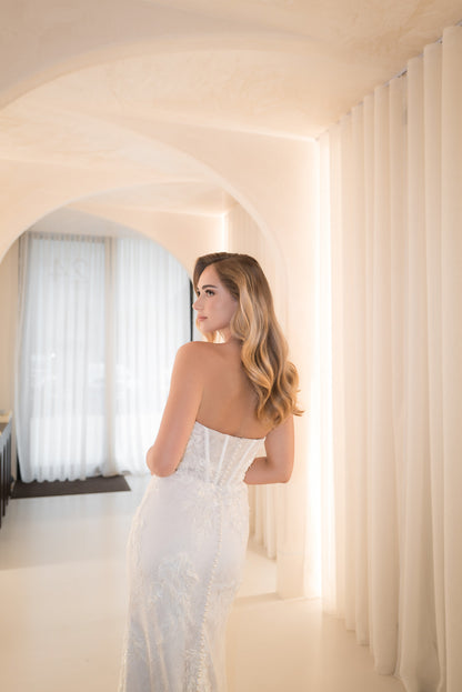 Woman in a white dress standing in a softly lit room with arches and curtains.