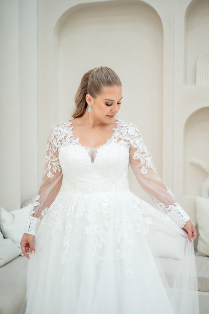 Woman in a white wedding dress with lace sleeves standing against a white wall.