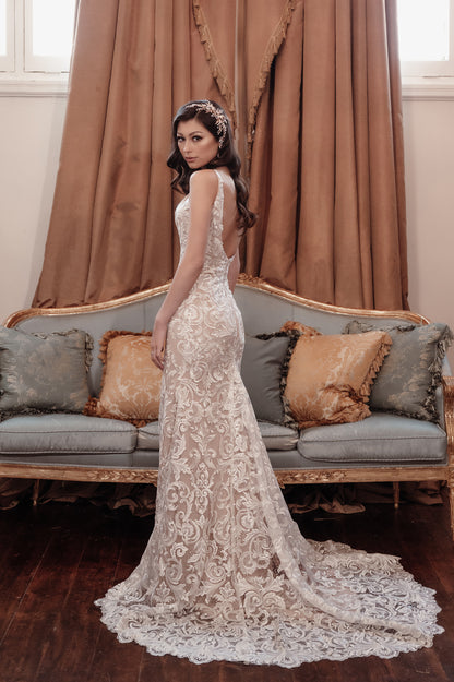 Woman in a white lace wedding dress standing in a room with a couch and decorative pillows.