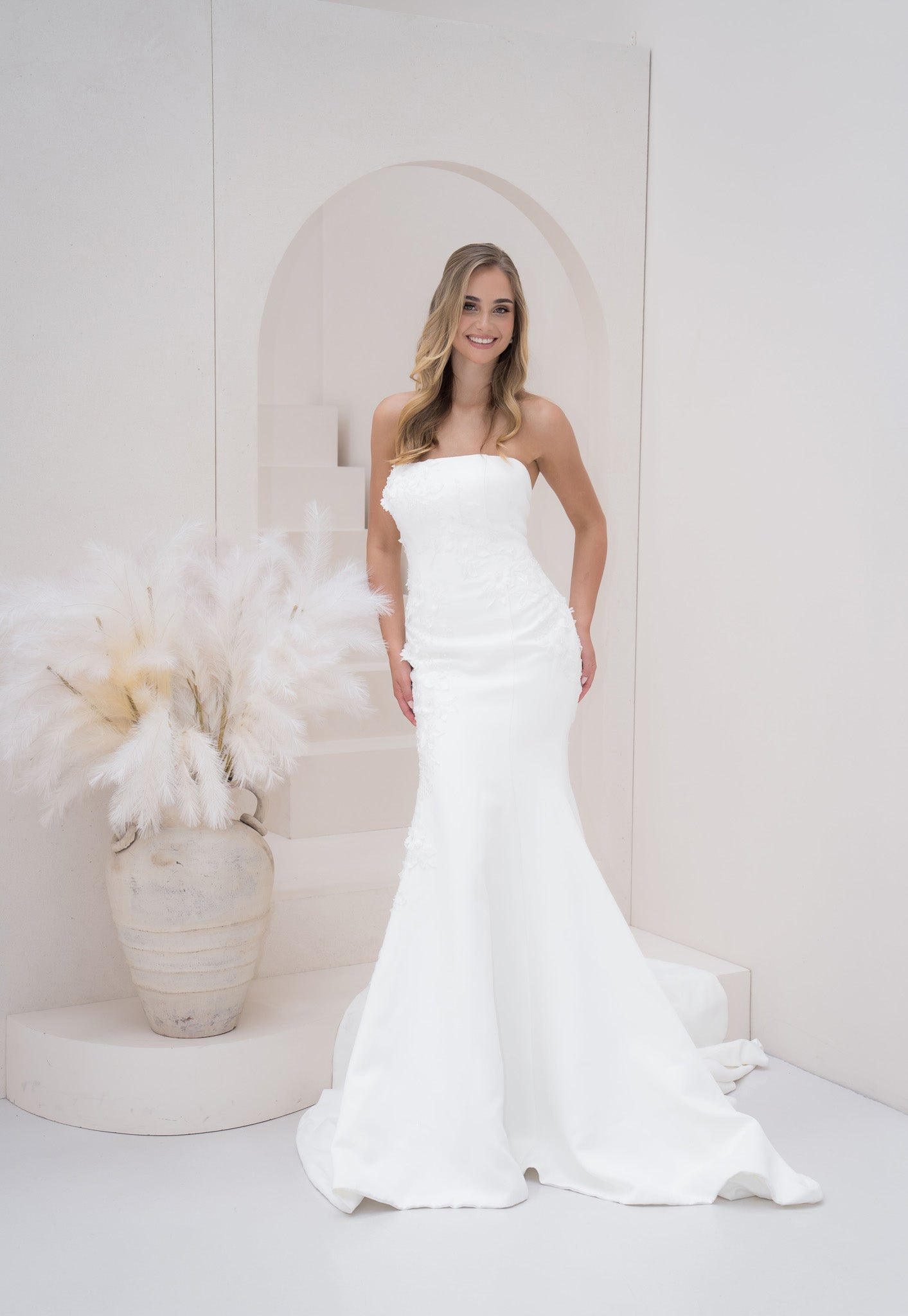 Woman in a white strapless gown standing in a minimalistic room with decorative elements.