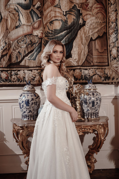 Woman in a white dress standing in front of ornate wall tapestries and decorative vases.