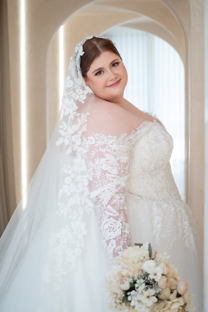 Woman in a white wedding dress with a veil holding a bouquet, standing in an archway.