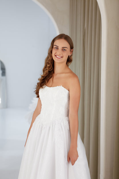 Woman wearing a white strapless dress standing in a softly lit room with neutral decor.