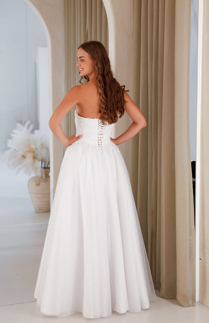 Woman in a white strapless gown standing in a room with beige curtains and decorative elements.