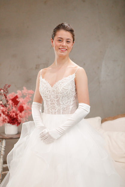 Woman in a white wedding dress with lace detailing and long gloves, sitting on a couch with a neutral background.